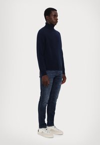 Navyblauer Rollkragenpullover aus weichem Strick, kombiniert mit dunklen Jeans und weißen Sneakern. Schlichtes und tailliertes Design.
