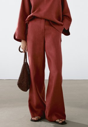 Personne portant un pantalon rouge ample et un top, tenant un sac à main marron perlé, et portant des sandales plates ouvertes à l'avant sur un sol gris.