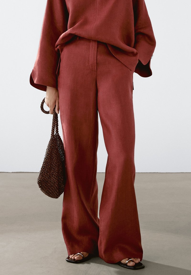 Personne portant un pantalon rouge ample et un top, tenant un sac à main marron perlé, et portant des sandales plates ouvertes à l'avant sur un sol gris.