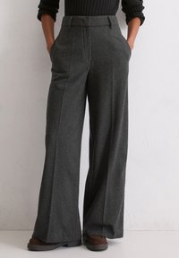 Pantaloni a gamba larga in tessuto grigio scuro con finitura texturizzata, caratterizzati da pieghe frontali e tasche laterali. Scarpe marroni parzialmente visibili.