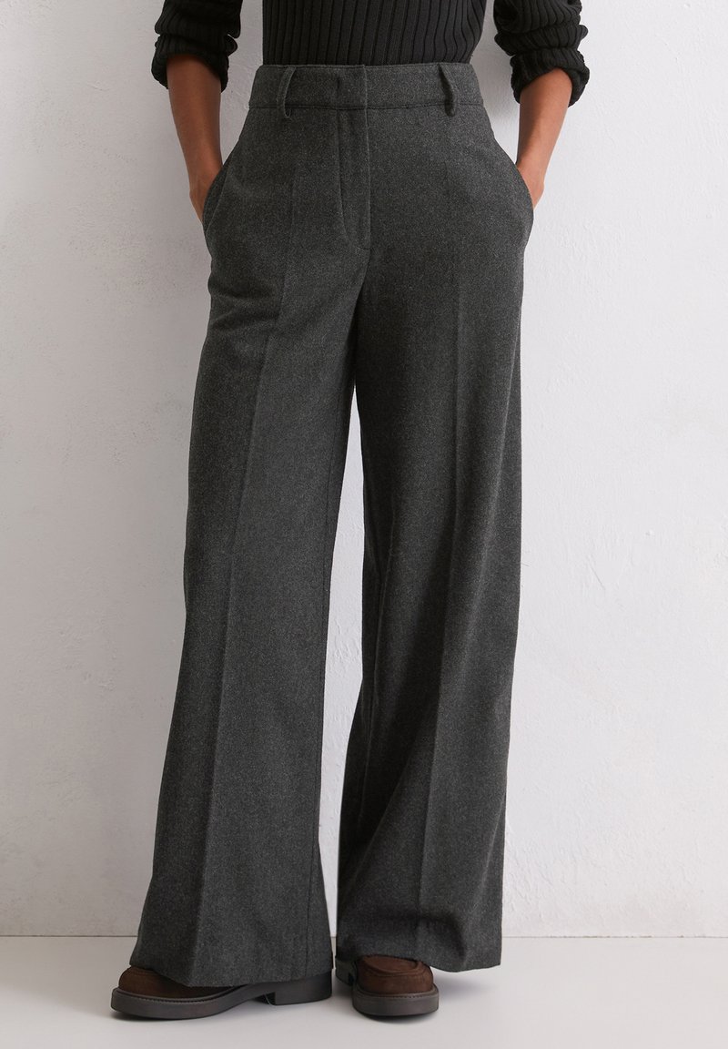Pantaloni a gamba larga in tessuto grigio scuro con finitura texturizzata, caratterizzati da pieghe frontali e tasche laterali. Scarpe marroni parzialmente visibili.