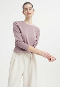 Helllila Langarmoberteil mit Rundhalsausschnitt, aus geripptem Stoff, kombiniert mit hochgeschnittenen cremefarbenen Hosen mit Falten.
