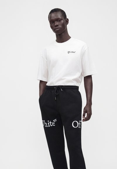 Μαύρος άνδρας στέκεται, φορώντας λευκό T-shirt Off-White και μαύρη φόρμα με μεγάλο κείμενο Off-White στα πόδια, σε απλό φόντο.