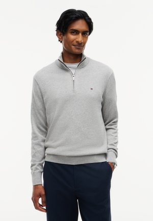 Homme portant un pull gris clair à fermeture éclair quart de longueur par-dessus une chemise rayée et un pantalon foncé, debout avec une main dans la poche sur fond blanc.