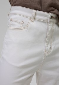 Jeans in denim bianco con un design attillato, cuciture visibili in marrone chiaro e una tasca laterale vicino alla vita. Texture liscia.