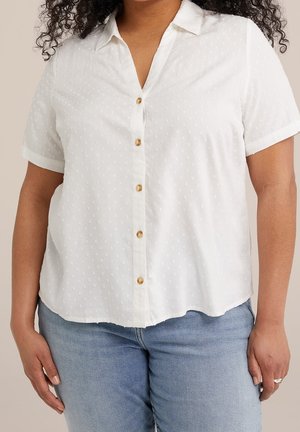 Femme portant un chemisier blanc à manches courtes, boutonné, avec une texture subtile à pois et un jean bleu clair, debout avec les bras détendus le long du corps.