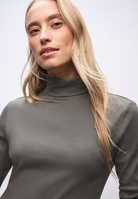 Maglione a collo alto grigio a costine con maniche lunghe, caratterizzato da un design aderente e una consistenza morbida, che si abbina ai toni naturali dei capelli e della pelle.