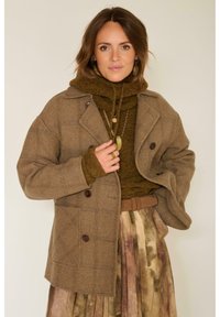 Manteau en laine à carreaux marron avec de gros boutons, superposé sur un pull en tricot vert foncé, associé à une jupe légère plissée multicolore.