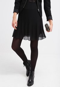 Jupe plissée noire avec des couches transparentes, portée avec des collants noirs et un haut en dentelle. Les accessoires comprennent une pochette métallique ronde et des bottines noires.