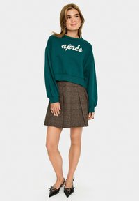 Groene cropped sweatshirt met witte tekst "après", gecombineerd met een bruine geruite geplooide rok en zwarte puntige hakken.