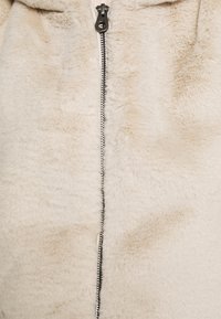 Krämfärgad konstpälsplagg med vertikal dragkedjestängning, med en mjuk textur och slät yta. Inga anmärkningsvärda mönster eller accenter.