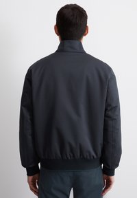 Tamsi, lengva bomber tipo striukė su aukšta apykakle, elastingomis rankovėmis ir apačia. Lygus paviršius ir minimalistinis dizainas, rodomas iš nugaros.