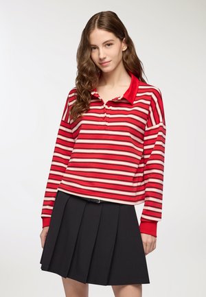 Polo a maniche lunghe a righe rosse e bianche con colletto rosso e placket dei bottoni, abbinato a una gonna nera a pieghe. Finitura del tessuto liscia.