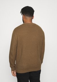 Hnědý pletený svetr se strukturovanou vazbou, kulatým výstřihem a žebrovanými manžetami, v kombinaci s černými kalhotami. Pohled zezadu.