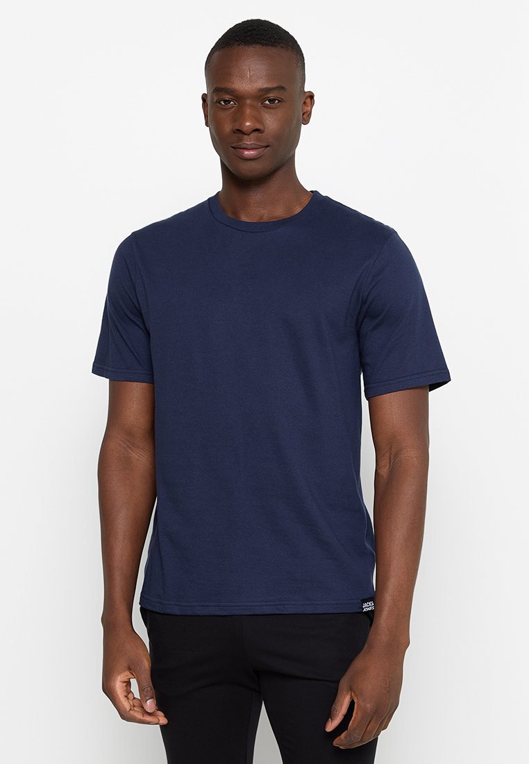 Joven hombre negro de pie mirando de frente, vistiendo una camiseta de manga corta azul marino lisa y pantalones negros, contra un fondo blanco.