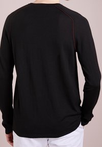 Persona che indossa una maglietta nera a maniche lunghe con cuciture rosse lungo le cuciture delle spalle e pantaloni bianchi, vista da dietro.