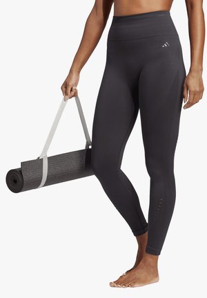 Leggings neri a vita alta con pannelli testurizzati e piccoli ritagli, abbinati a un tappetino da yoga nero arrotolato fissato da una cintura grigia chiara.