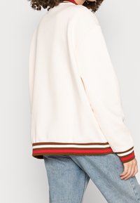 Giacca bomber color crema con vestibilità rilassata, polsini a coste e orlo a righe con accenti rossi, marroni e bianchi. Tessuto morbido.
