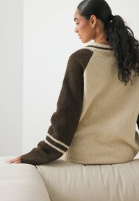 Krēmkrāsas un brūnas adītas džemperis ar raglan stilu, kura piedurknēm ir baltas svītras. Mīksta tekstūra, brīvs griezums un kontrastējošas krāsas.