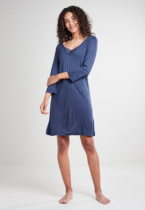 Frau mit lockigem Haar, die ein langärmeliges dunkelblaues Kleid mit Spitzenbesatz trägt und barfuß vor einem schlichten weißen Hintergrund steht.