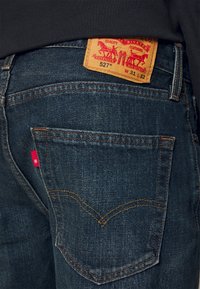 Jeans de mezclilla oscura con un ajuste clásico, que cuentan con un parche de cuero en la parte trasera con la marca y una etiqueta roja en el bolsillo trasero.