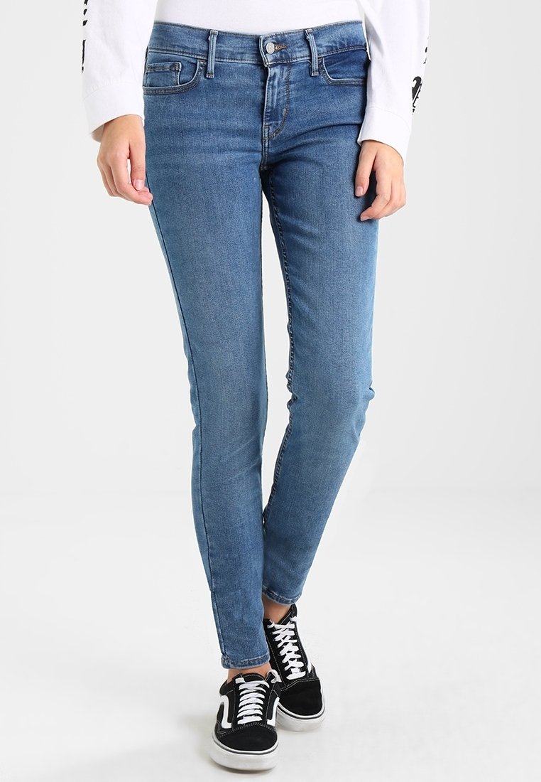 Jeans de mezclilla azul ajustados con un lavado medio, cinco bolsillos y un cierre de botón clásico. Se llevan con zapatillas negras que tienen cordones blancos.