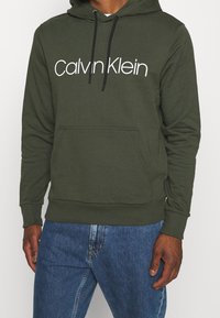 Bluza z kapturem w oliwkowej zieleni z przednią kieszenią, czarnymi sznurkami i białym napisem "Calvin Klein" na piersi. Regularny krój.