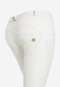 Pantaloni bianchi aderenti con una texture liscia, dotati di tasche posteriori e un piccolo emblema dorato. Progettati con una silhouette su misura.