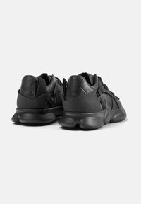 Schwarze Sportschuhe mit einer strukturierten Oberfläche, gepolstertem Absatz und einer auffälligen Sohle mit wellenförmigem Design. Schnürsenkel und elastische Bänder für einen sicheren Sitz.