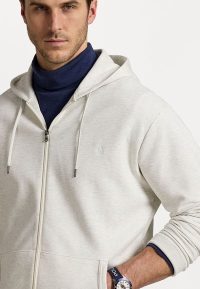 Light gray zip-up hoodie with a textured fabric, featuring a drawstring hood and a subtle embroidered logo on the chest. Blue turtleneck underneath.