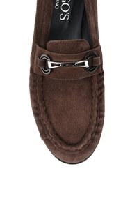 Brązowe zamszowe mokasyny z detalem w postaci srebrnej klamry na górze i czarną wkładką z częściowo widocznym białym tekstem.