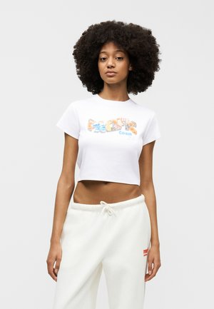 Νεαρή γυναίκα με σγουρά μαλλιά, που φοράει λευκό κοντομάνικο μπλουζάκι με τύπωμα και λευκό παντελόνι με κορδόνι, μπροστά σε απλό φόντο.