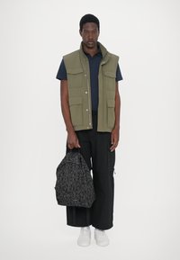 Grön ärmlös väst med flera fickor över en marinblå t-shirt, kombinerad med svarta cargo-byxor och en svart mönstrad ryggsäck. Vita sneakers.