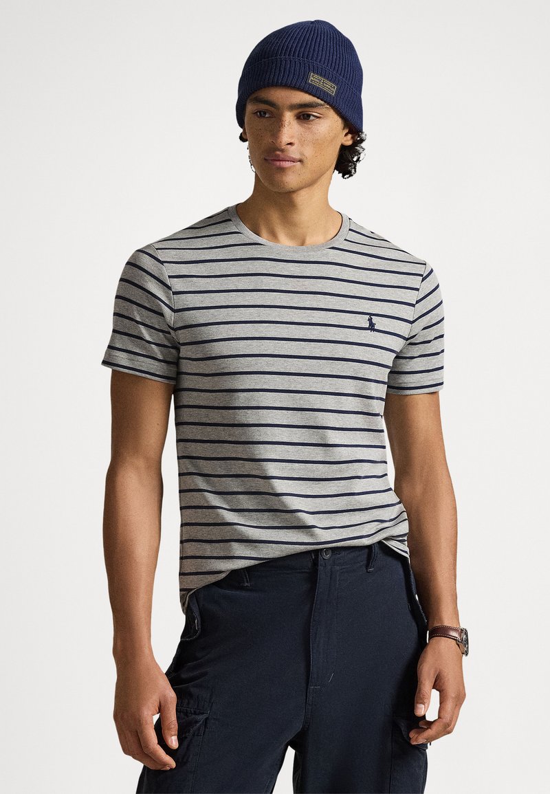 Grijs gestreept T-shirt met marineblauwe horizontale lijnen, voorzien van een ronde hals en een klein logo op de borst, gecombineerd met een donkere broek en een blauwe muts.