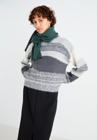 Maglione a righe grigio, bianco e crema con una texture morbida, abbinato a una sciarpa rigata verde, indossato su una gonna nera.