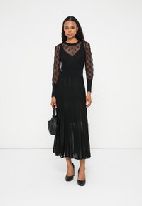 Vestido negro con mangas largas y transparentes, con estampado, cuerpo ajustado y falda plisada y fluida. Combinado con tacones negros y un bolso pequeño.