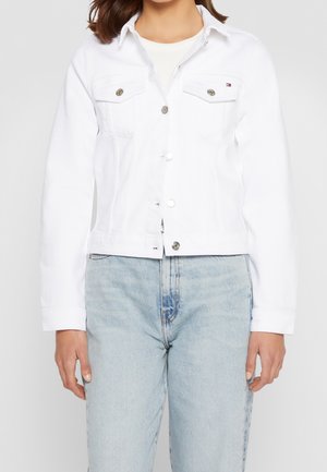 Kvinde iført hvid denimjakke med sølvknapper og lyseblå højtaljede jeans, stående mod ensfarvet baggrund.