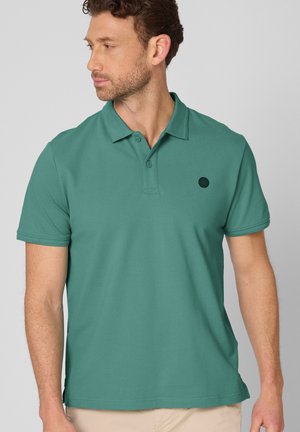 Mężczyzna w zielonym polo z krótkim rękawem z dwoma guzikami, patrzący w lewo, ubrany w beżowe spodnie na jednolitym jasnym tle.