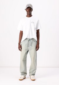 Hvid t-shirt med et lille grafisk motiv, lyseblå jeans med elastisk snøre i taljen, grå kasket med farverige detaljer og hvide sneakers.