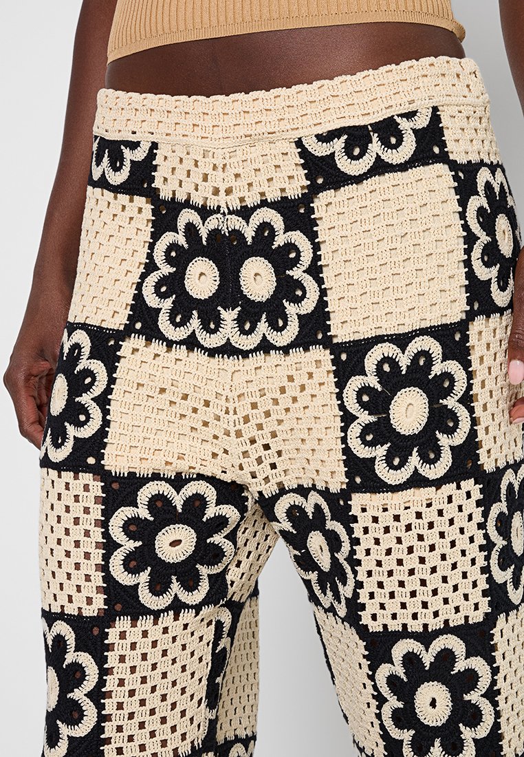 Hohe gehäkelte Hosen mit schwarz-beigen Patchwork-Quadraten, die Blumens- und Rautenmuster zeigen, getragen mit einem kurzen beigen Oberteil.