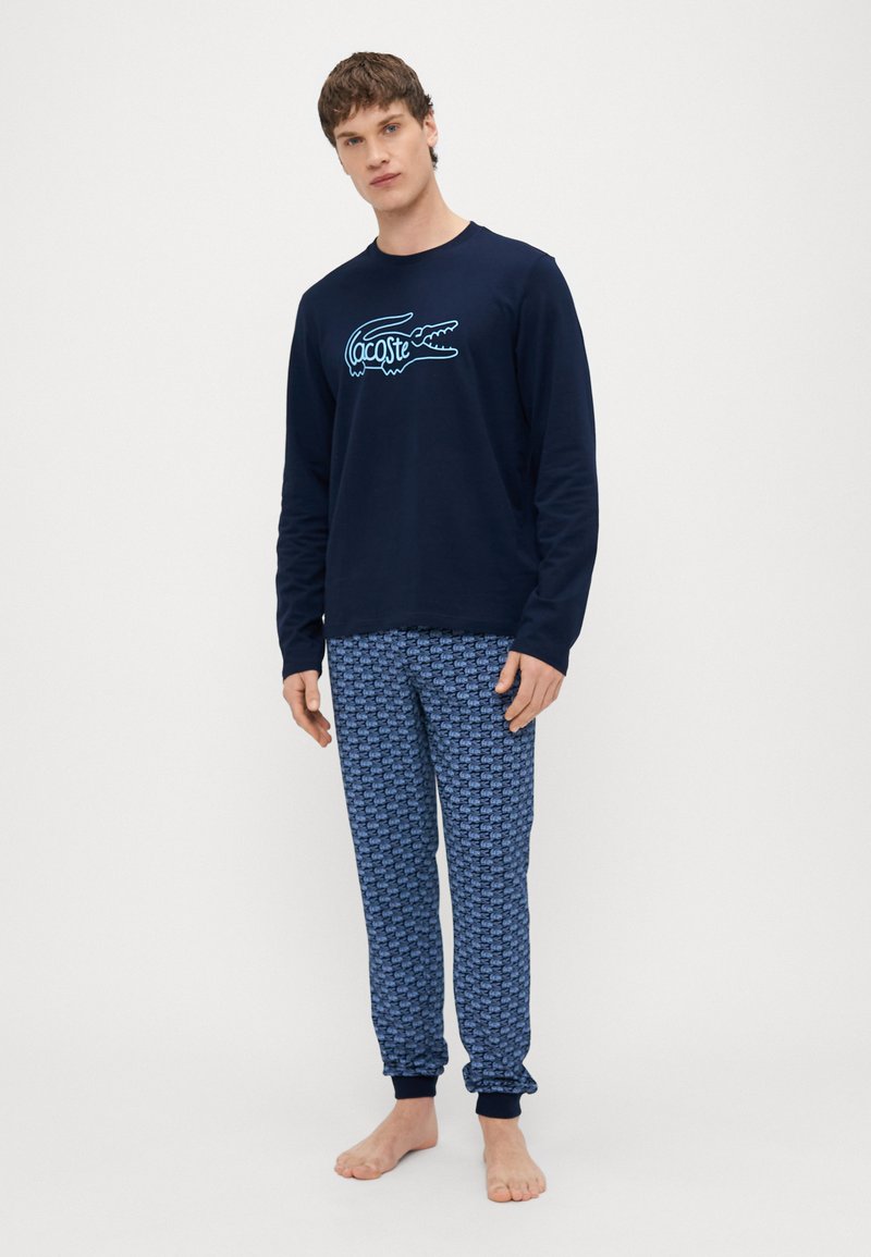 Top de manga larga azul marino con el logotipo de cocodrilo "Lacoste" en azul claro, combinado con pantalones de pijama azules con un patrón repetido de logotipos.