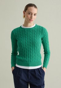 Maglione verde a treccia con orlo e polsini a costine, indossato su una camicia bianca a girocollo. Il modello indossa pantaloni blu navy plissettati.