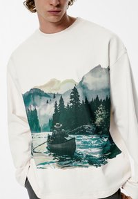 Světlý mikina v barvě off-white s akvarelovým motivem rybáře v kánoi mezi horami a stromy, s měkkou, texturovanou látkou.