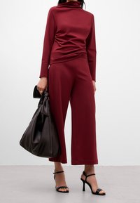 Top bordeaux a maniche lunghe con collo alto e design drappeggiato, abbinato a pantaloni a gamba larga. Completato da una grande borsa marrone scuro e tacchi neri.