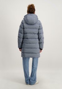 Grijze pufferjas met een afneembare capuchon. Heeft gequiltte secties, lange mouwen en zwarte accenten. Gepaard met blauwe jeans en witte sneakers.