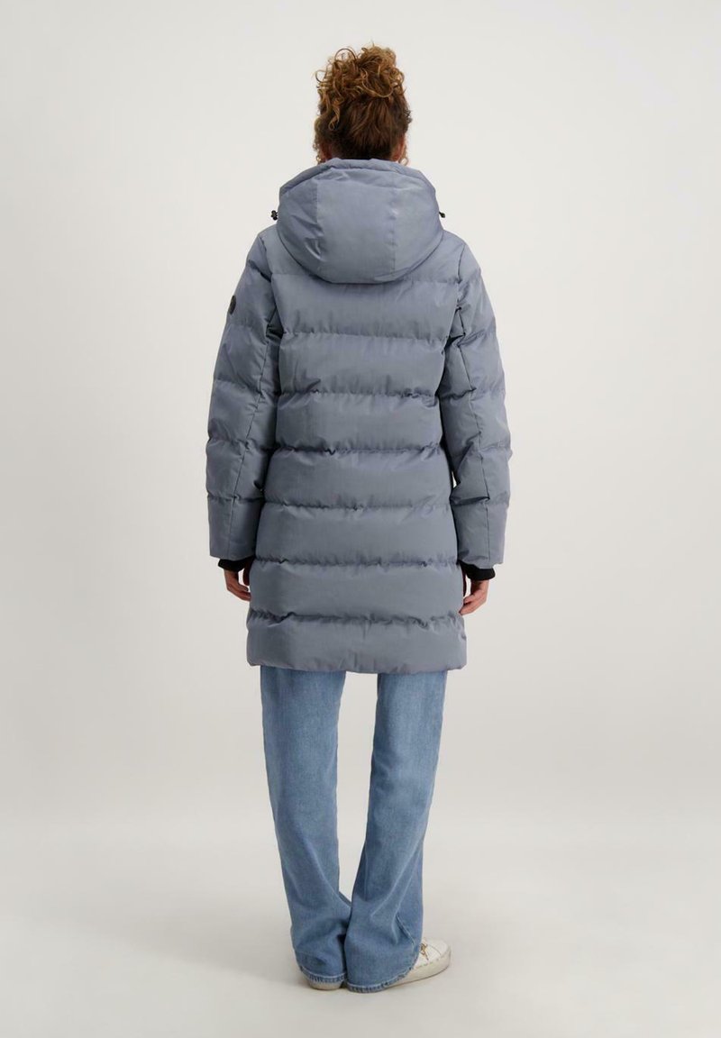 Grijze pufferjas met een afneembare capuchon. Heeft gequiltte secties, lange mouwen en zwarte accenten. Gepaard met blauwe jeans en witte sneakers.