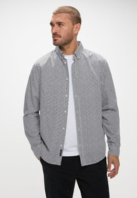 Camisa de botones en un patrón geométrico en blanco y negro, con cuello extendido, mangas largas y dobladillo recto. Llevada sobre una camiseta blanca.