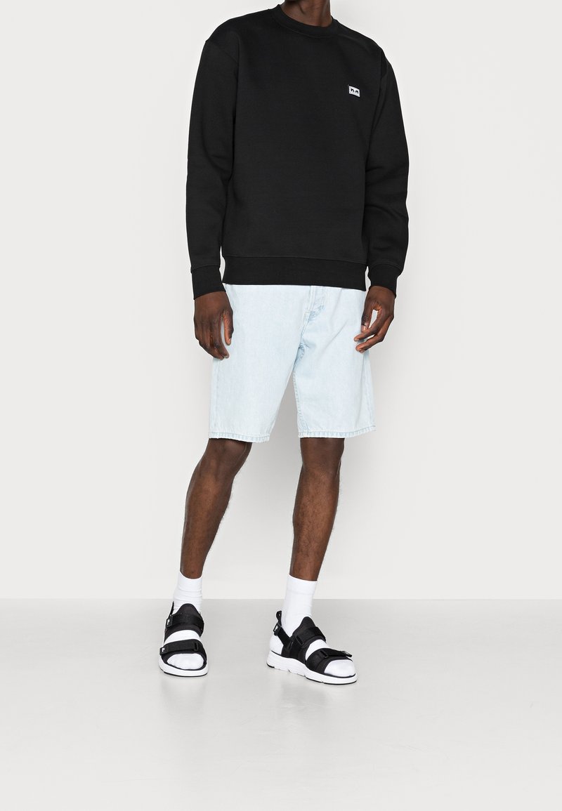 Personne portant un sweat-shirt noir à manches longues, un short en denim bleu clair arrivant au genou, des chaussettes blanches et des sandales noires sur un fond clair uni.