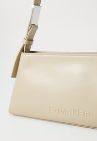 Bež torba od kože s glatkocrnom teksturom, vitkim dizajnom i ugraviranim logotipom Calvin Klein. Ima odvojivu naramenicu.