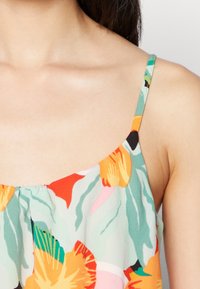 Bloemenprint top met verstelbare dunne sp straps. Het materiaal lijkt lichtgewicht en vloeiend, met levendige kleuren zoals groen, oranje en roze.